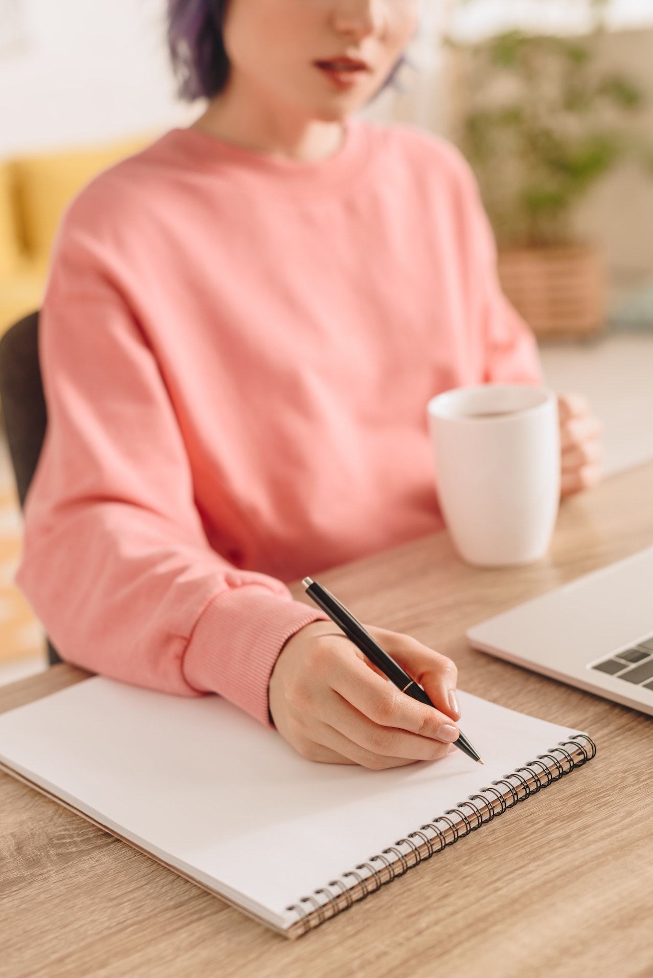 Cropped view of freelancer holding cup of tea and writing in notebook at table